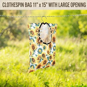May include: A clothes pin bag, 11" x 15", with a large opening, hanging on a clothesline. The bag is decorated with a sunflower pattern in yellow, green, and white. The bag has a black trim and wooden clothes pins.