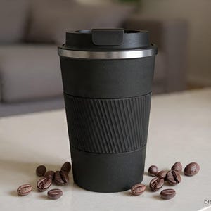 May include: A black travel mug with a silver rim and a textured grip. Coffee beans are scattered around the base of the mug. The mug is designed to keep beverages hot or cold. The mug is approximately 7 inches tall.