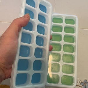 May include: Two rectangular ice cube trays. One tray is filled with blue ice cubes, and the other is filled with green ice cubes. The trays have a white frame and are made of a flexible material.