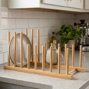 May include: A natural bamboo dish rack with vertical dowels to hold plates and mugs. A beige plate and a ceramic mug are placed in the rack. The rack is on a light gray countertop in a kitchen setting.