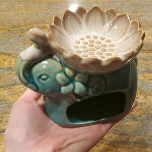 May include: A ceramic oil burner shaped like an elephant with a teal crackle glaze finish. The elephant supports a large, beige, lotus-shaped bowl on its back, featuring a textured center with small raised dots for holding wax or oil.