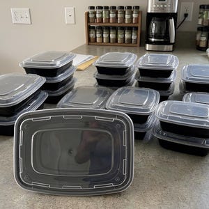 May include: A collection of rectangular black plastic food storage containers with clear, snap-on lids. The containers are arranged in multiple stacks of varying heights, with one single container positioned in the foreground showing the lid detail.