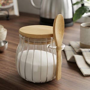 May include: A clear glass jar with vertical ribbed detailing, featuring a round light-colored wooden lid and a matching wooden spoon held in a small glass loop on the side. The jar is filled with white granulated sugar.