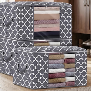 May include: Two rectangular storage bins with a dark gray and white quatrefoil pattern. Each bin features a clear plastic front window revealing folded clothing inside, a zippered closure, and two gray fabric handles on the sides.