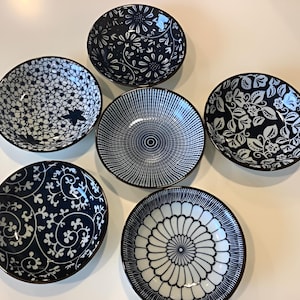 May include: A set of six round ceramic bowls featuring various dark blue and white patterns. The designs include floral motifs, geometric lines, a star, and swirling vines. Each bowl has a dark rim and a smooth, circular shape for serving.