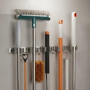 May include: A wall-mounted stainless steel organizer rack featuring multiple spring-loaded slots and hooks. It holds various cleaning tools including a broom with a teal head, a brown duster, and several long-handled mops and brushes with orange and white accents.