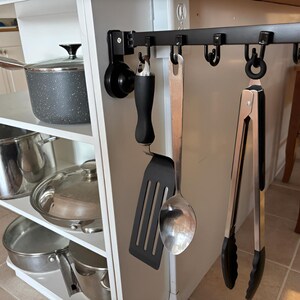 May include: A white kitchen island with stainless steel pots and pans on shelves. A black metal rack holds cooking utensils, including a spatula, spoon, and tongs. The pots and pans are various sizes and have lids.