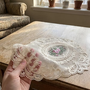 May include: An off-white, oval-shaped lace doily with an embroidered floral center featuring pink and green flowers. The doily has intricate lace detailing around the edges. The doily is held up, showcasing the delicate lace design.