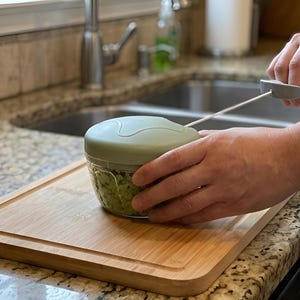 May include: A manual food chopper with a sage green lid and a clear plastic bowl containing chopped green vegetables. The device features a pull-cord mechanism with a grey handle, designed for slicing ingredients through a hand-operated motion.