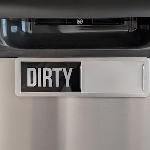 May include: A rectangular dishwasher magnet with a sliding mechanism, featuring a black panel with the white text DIRTY on the left side and a blank silver panel on the right, mounted on a stainless steel dishwasher door with control settings.