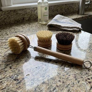 May include: Three wooden dish brushes with natural bristles. One brush has a long wooden handle with a metal loop. The other two brushes are round with wooden bases. A bottle of dish soap and a folded dish towel are in the background.