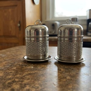 May include: Two cylindrical stainless steel shakers with a polished finish and a repeating hexagonal honeycomb pattern etched into the lower section. Each shaker features a rounded dome top with a small metal loop handle and sits on a matching base.