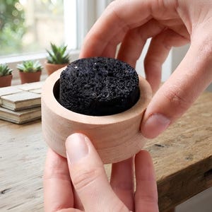 May include: A cylindrical light-colored wooden container holding a circular, textured, porous black charcoal disc. The container has a smooth, unfinished wood grain surface and a simple, open-top design that frames the dark, rough-surfaced insert.