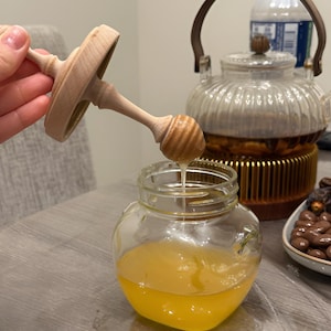 May include: A clear glass jar filled with golden honey, accompanied by a wooden honey dipper with a matching lid. The dipper is positioned above the open jar, with a thin stream of honey dripping from the grooved head into the container below.