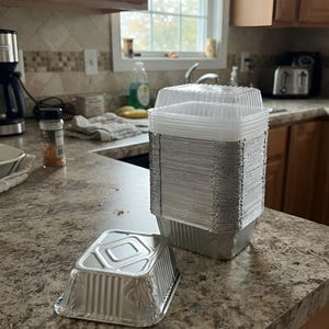May include: A stack of square aluminum foil food containers with clear plastic lids, accompanied by a single separate aluminum foil container placed in front. The containers feature a ribbed side design and a flat, reinforced base for stability.