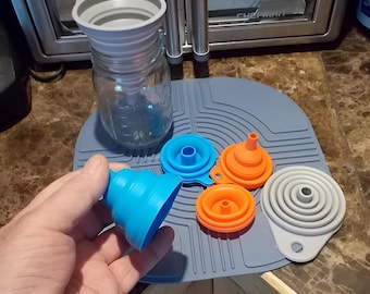 May include: A set of collapsible silicone funnels in blue, orange, and gray, featuring a wide mouth and a narrow spout. One gray funnel is inserted into a clear glass jar, while the others are arranged on a gray silicone mat with ridges.