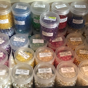 May include: A collection of clear plastic jars filled with various colors of beads, including yellow, blue, green, purple, red, and gold. The jars are labeled with bead size and material, such as "8 mm Resin" and "12 mm Resin".