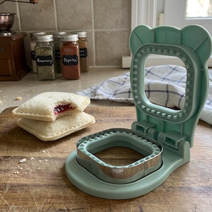 May include: A light green plastic sandwich sealer with a bear-shaped top and a metal square cutter base. Next to the tool are two square white bread sandwiches with crimped edges, one showing a red jam filling inside the center of the bread.