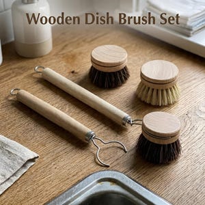 May include: Wooden dish brush set with three round brushes and two wooden handles. The brushes have light brown wooden bases and dark bristles. The handles are light wood with metal loops. The text "Wooden Dish Brush Set" is at the top.