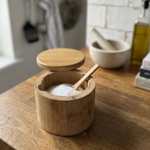 May include: A cylindrical bamboo salt cellar with a swivel lid and a small wooden spoon resting inside. The container is filled with white salt crystals and features a magnetic closure mechanism on the side of the rim for the lid.