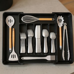 May include: A black plastic utensil organizer filled with various kitchen tools. Includes a slotted spoon, spatula, whisk, tongs, forks, knives, spoons, and a basting brush. The utensils have wooden or silver handles.