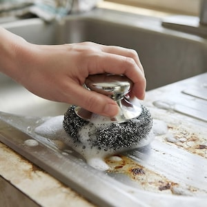 May include: A stainless steel pot scrubber featuring a rounded, polished metal handle and a dense, coiled wire mesh base. The scrubber is covered in white soap suds and is positioned on a metallic surface with visible dark residue and stains.