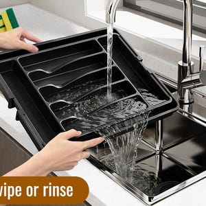 Puede incluir: Una bandeja negra para cubiertos con m&uacute;ltiples compartimentos se sostiene sobre un fregadero de acero inoxidable. El agua fluye de un grifo a la bandeja. El texto "Easy to wipe or rinse" se muestra en un cuadro marr&oacute;n en la parte inferior de la imagen.