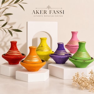 Könnte beinhalten: Eine Sammlung farbenfroher marokkanischer Tajine-Töpfe. Jede Tajine hat eine einzigartige Farbe, darunter Rot, Orange, Gelb, Lila, Rosa und Grün, mit goldfarbenen Streifen. Die Tajines werden auf weißen Sockeln präsentiert.