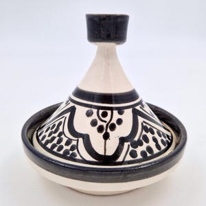 Puede incluir: Una olla de tagine de cerámica con tapa cónica y base redonda. La olla es principalmente blanquecina con patrones y acentos decorativos negros. La tapa tiene un asa negra y el borde también es negro.