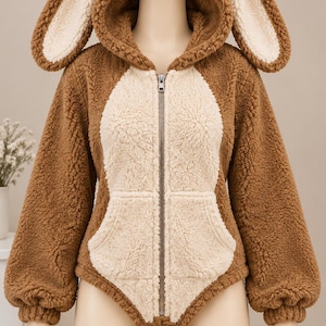 Puede incluir: Una sudadera con capucha y cremallera en color marrón y crema, confeccionada en tejido polar suave. La prenda cuenta con mangas largas con puños acanalados, cierre de cremallera frontal y una capucha con dos grandes orejas de conejo erguidas.