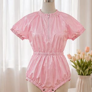 May include: A light pink short-sleeved romper made of a shiny, vinyl-like material. The garment features a high neckline with a central vertical zip, an elasticated waist, and elasticated leg openings with press stud closures at the bottom crotch area.