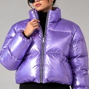 Puede incluir: Una chaqueta acolchada de color morado metálico brillante con cuello alto y cremallera frontal plateada. La prenda presenta paneles acolchados horizontales, mangas largas con puños ajustados y un corte corto a la cintura.