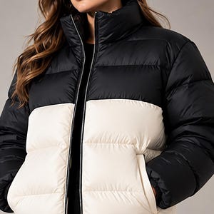 May include: A color-blocked puffer jacket featuring a black upper section and a cream-colored lower section. The jacket has a high collar, a full-length front zipper, and two side pockets. The design includes horizontal quilted stitching throughout.