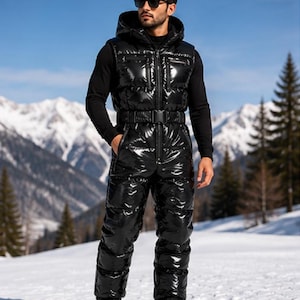 Puede incluir: Un mono de nieve acolchado de color negro con acabado brillante, que presenta una parte superior sin mangas con capucha, cierre de cremallera frontal y un cinturón con hebilla de plástico. Se lleva sobre una camiseta negra de manga larga.