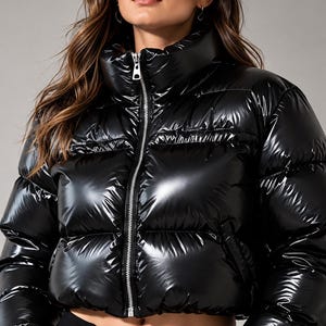 Puede incluir: Una chaqueta acolchada negra de alto brillo con silueta corta, cuello alto y cremallera frontal plateada de longitud completa. La prenda presenta canales acolchados horizontales y mangas largas con puños elásticos, ofreciendo un acabado brillante.