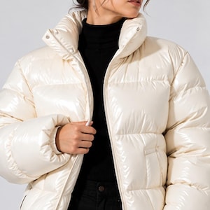 May include: A glossy cream-colored puffer jacket with a high collar, a full-length front zipper, and horizontal quilted panels. The jacket features long sleeves with elasticized cuffs and two side pockets, worn over a black mock neck top.