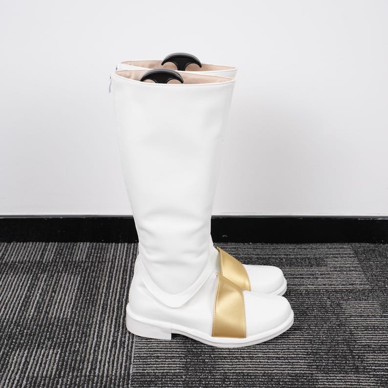 May include: White knee-high boots with a gold-colored strap across the foot. The boots have a smooth, clean finish and a low heel. The top of the boots have black handles. The boots are made of a synthetic material.