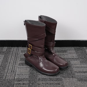 Może przedstawiać: Para brązowych butów z błyszczącym wykończeniem. Buty mają wysokość do połowy łydki, z paskami i złotymi klamrami. Podeszwy są czarne, a buty są ustawione na neutralnym tle.