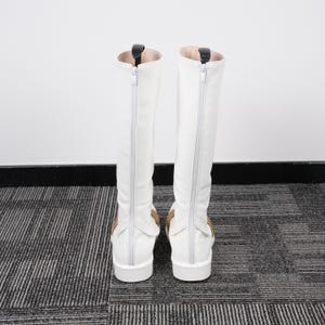 May include: A pair of white knee-high boots with a zipper down the back. The boots have a small heel and a tan accent near the ankle. The interior of the boot is a light tan color.