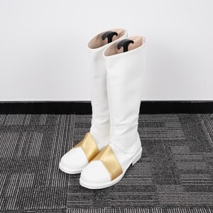 May include: A pair of white knee-high boots with gold accents on the foot. The boots have a smooth, clean design and are standing upright. Black boot shapers are inside the boots.