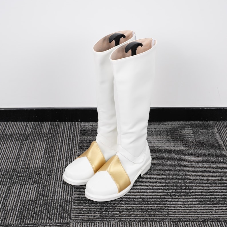May include: A pair of white knee-high boots with gold accents on the foot. The boots have a smooth, clean design and are standing upright. Black boot shapers are inside the boots.
