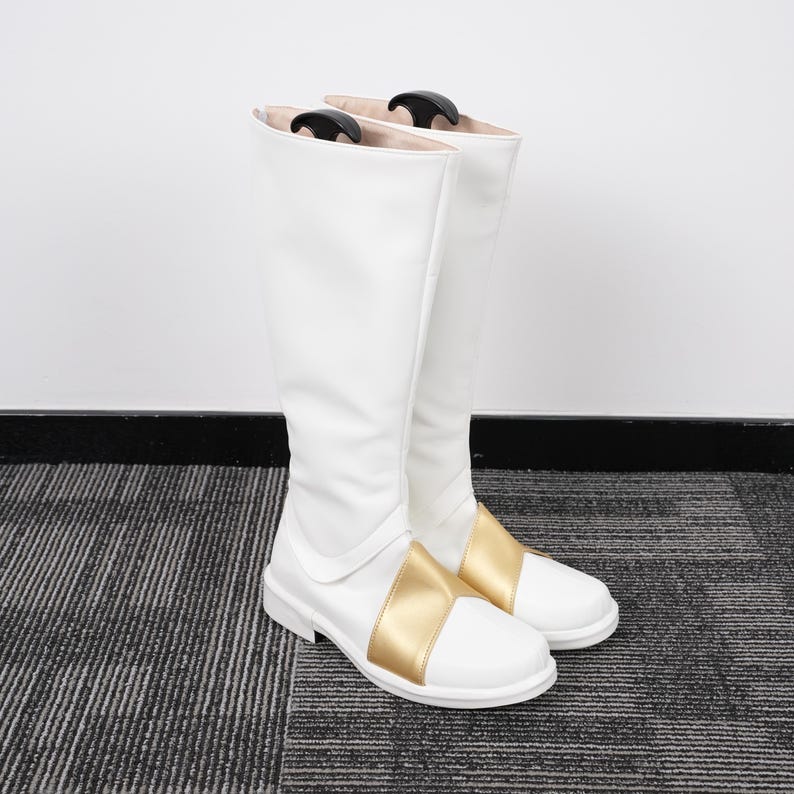 May include: Pair of white knee-high boots with a gold band across the foot. The boots have a smooth, glossy finish and a simple, elegant design. The boots are standing upright on a gray and black striped surface.