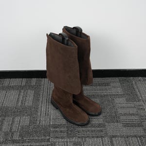 Pode incluir: Um par de botas de camurça marrom com um design até o joelho. As botas têm uma parte superior dobrada e uma sola preta. As botas são exibidas em um tapete estampado contra uma parede branca.
