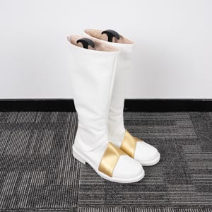 May include: A pair of white knee-high boots with a gold band across the foot. The boots are made of a smooth material and have a slight sheen. Black boot shapers are inside the boots.