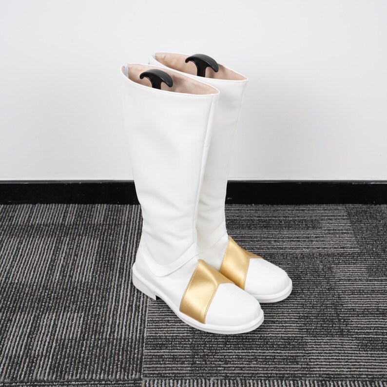 May include: A pair of white knee-high boots with a gold band across the foot. The boots are made of a smooth material and have a slight sheen. Black boot shapers are inside the boots.
