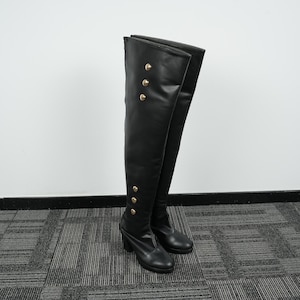 May include: A pair of black over-the-knee boots. The boots are made of a smooth material and feature gold-colored buttons down the side. The boots have a block heel and a rounded toe. The boots are standing on a gray and black carpet.