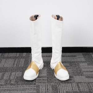 Puede incluir: Un par de botas blancas hasta la rodilla con un acento dorado en el pie. Las botas tienen una cremallera en la parte trasera y están hechas de un material liso, probablemente sintético. Las botas están de pie contra una pared blanca.