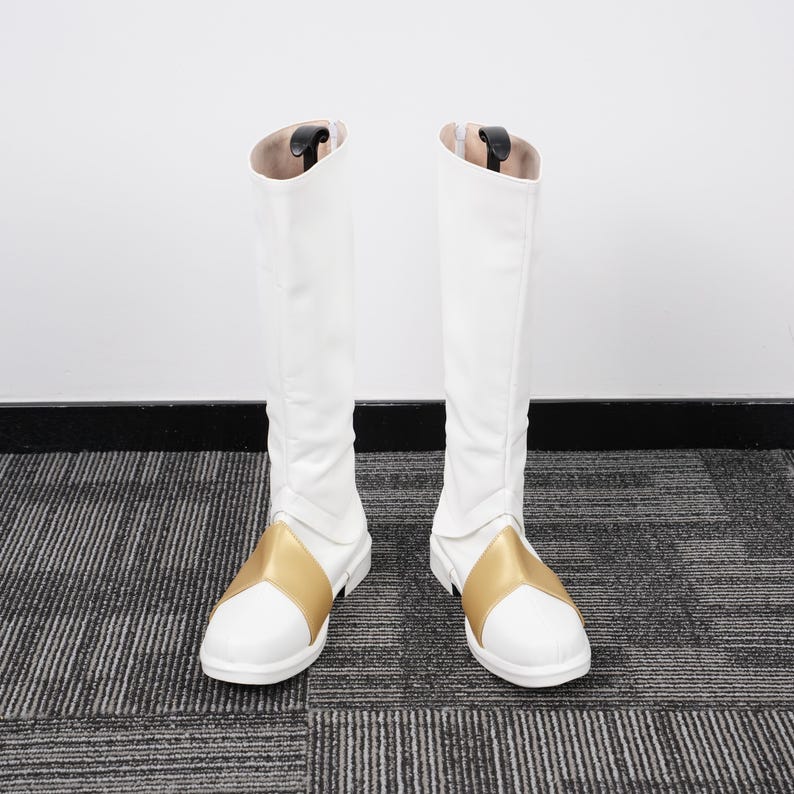 May include: A pair of white knee-high boots with a gold accent on the foot. The boots have a zipper on the back and are made of a smooth, likely synthetic material. The boots are standing upright against a white wall.