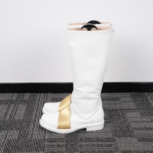 May include: A pair of white knee-high boots with a gold-colored strap across the front. The boots have a smooth, glossy finish and a low heel. The boots are designed for costume or cosplay.