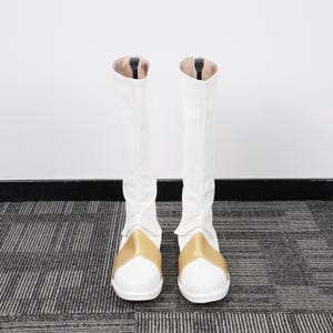 May include: A pair of white knee-high boots with gold accents. The boots have a smooth, clean design with a gold band across the top of the foot. The interior of the boots is a light tan color.